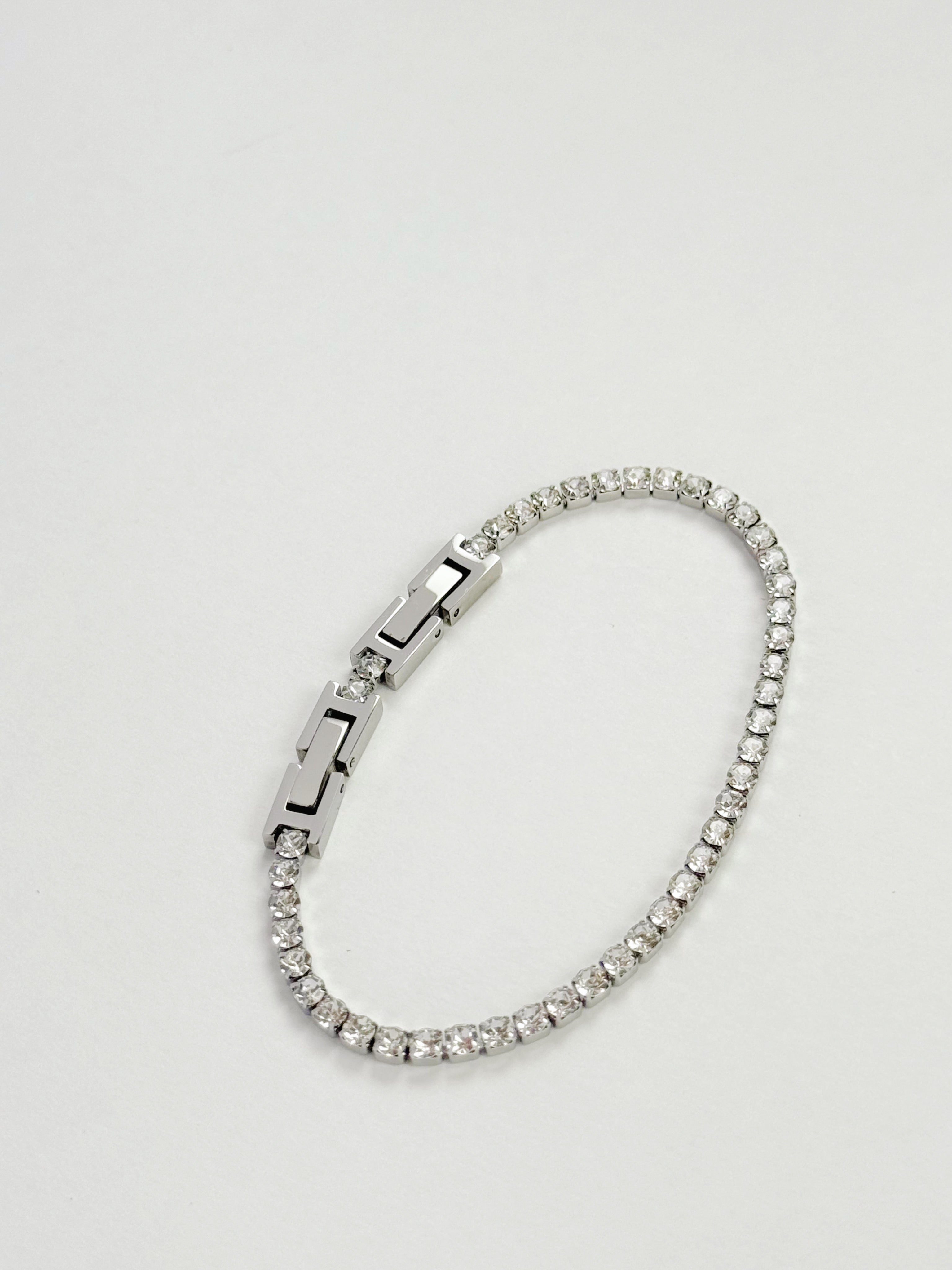 A silver-toned tennis bracelet with clear stones and a geometric clasp, photographed on a light gray background.