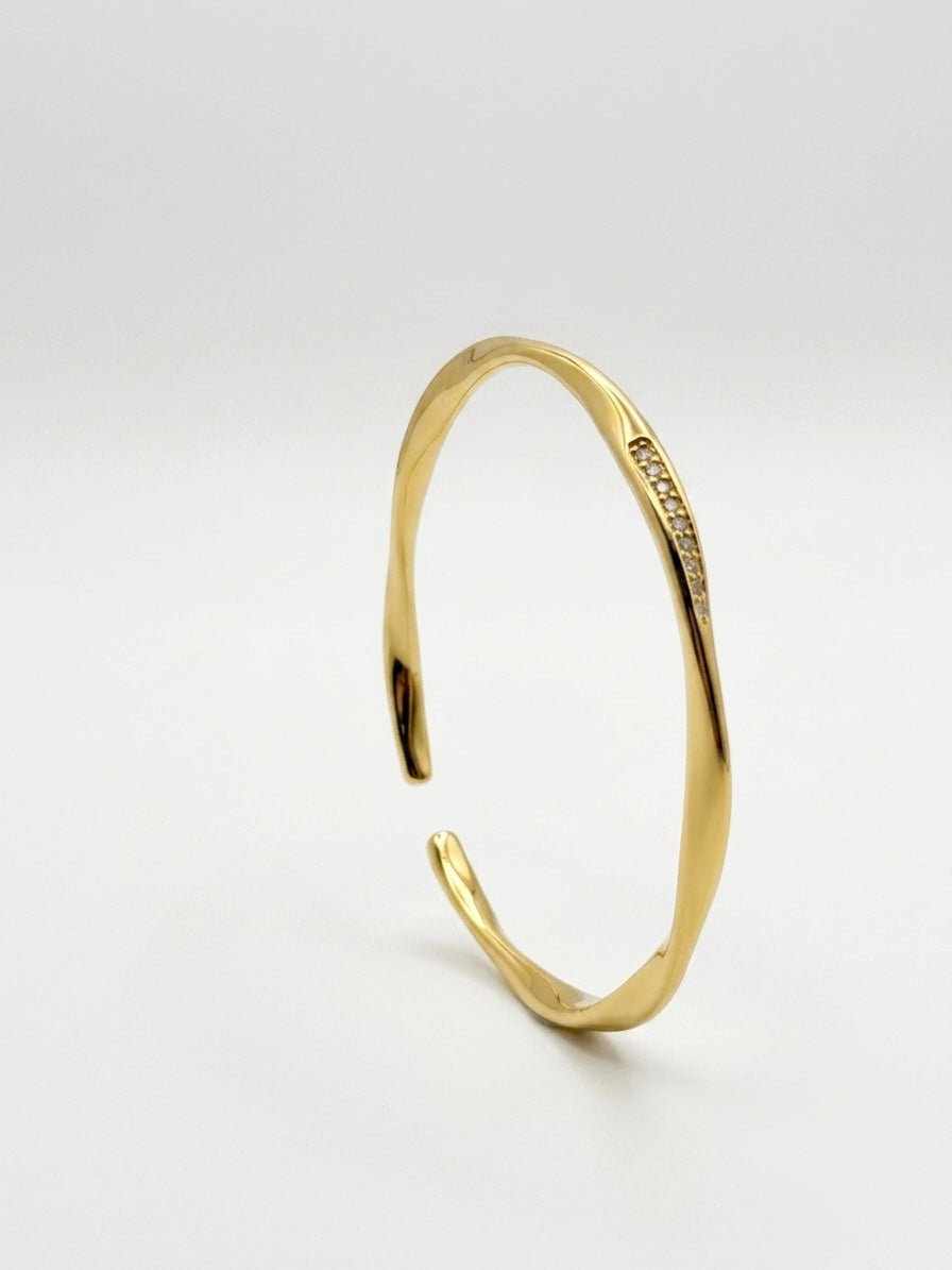 A polished gold-tone open cuff bangle bracelet with a small line of clear stones set along one side, photographed on a seamless light gray studio background.