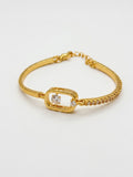 A gold bangle bracelet featuring a central open rectangular frame with a clear round gemstone, flanked by small pavé-set diamonds and a delicate chain extender clasp, shown on a plain white background.