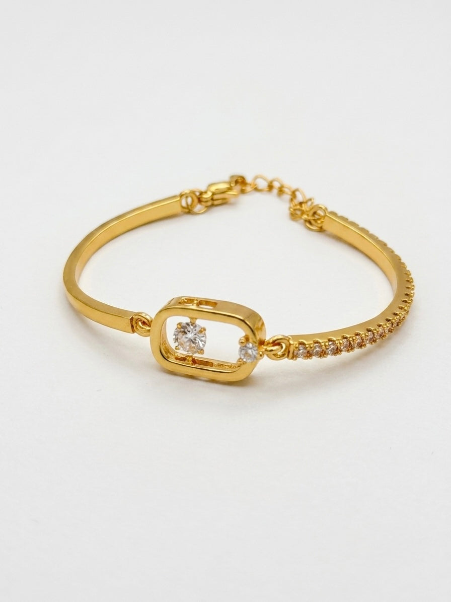 A gold bangle bracelet featuring a central open rectangular frame with a clear round gemstone, flanked by small pavé-set diamonds and a delicate chain extender clasp, shown on a plain white background.