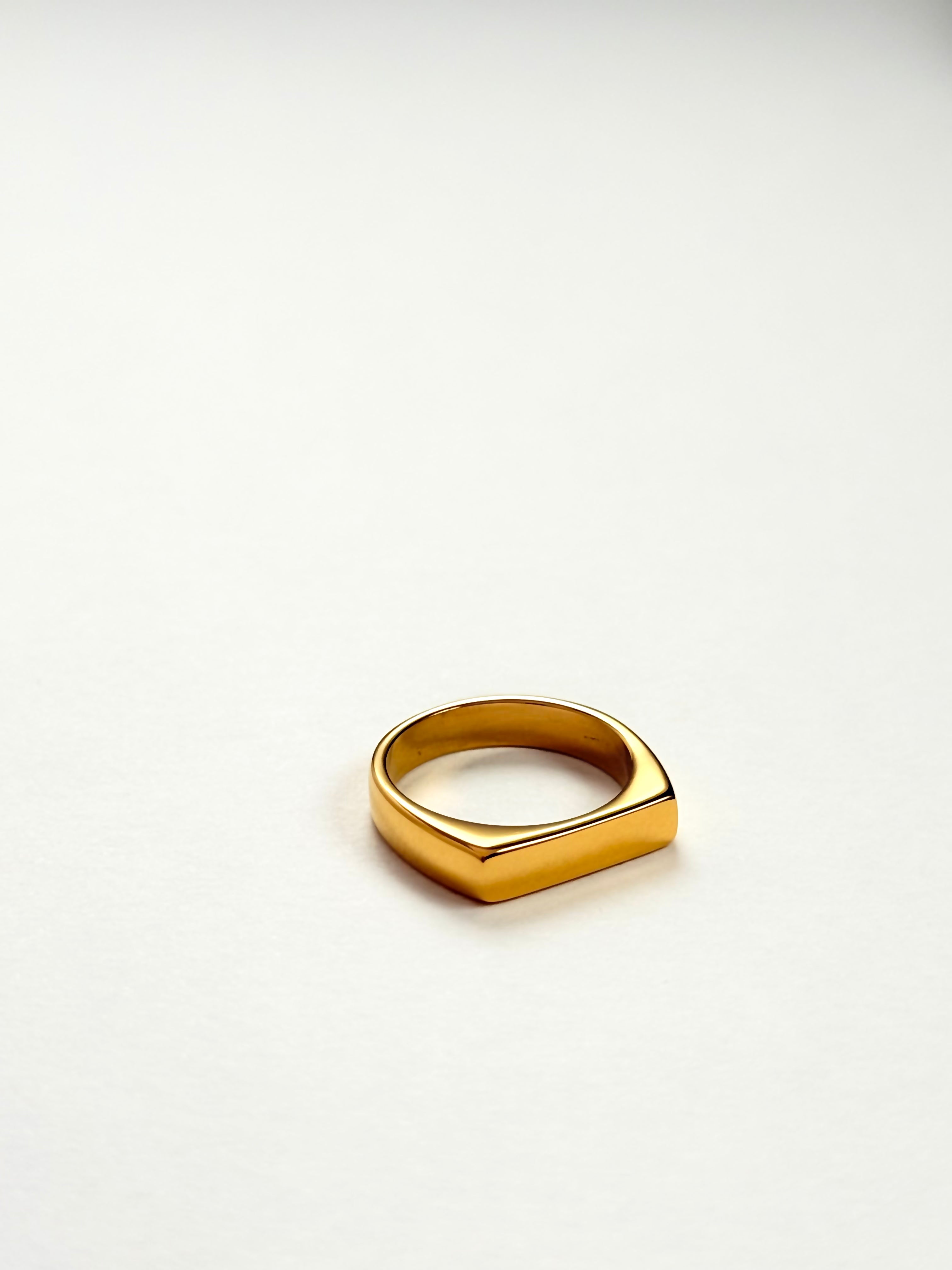 A polished gold signet ring with a geometric, slightly angled square face, resting on a clean white surface.
