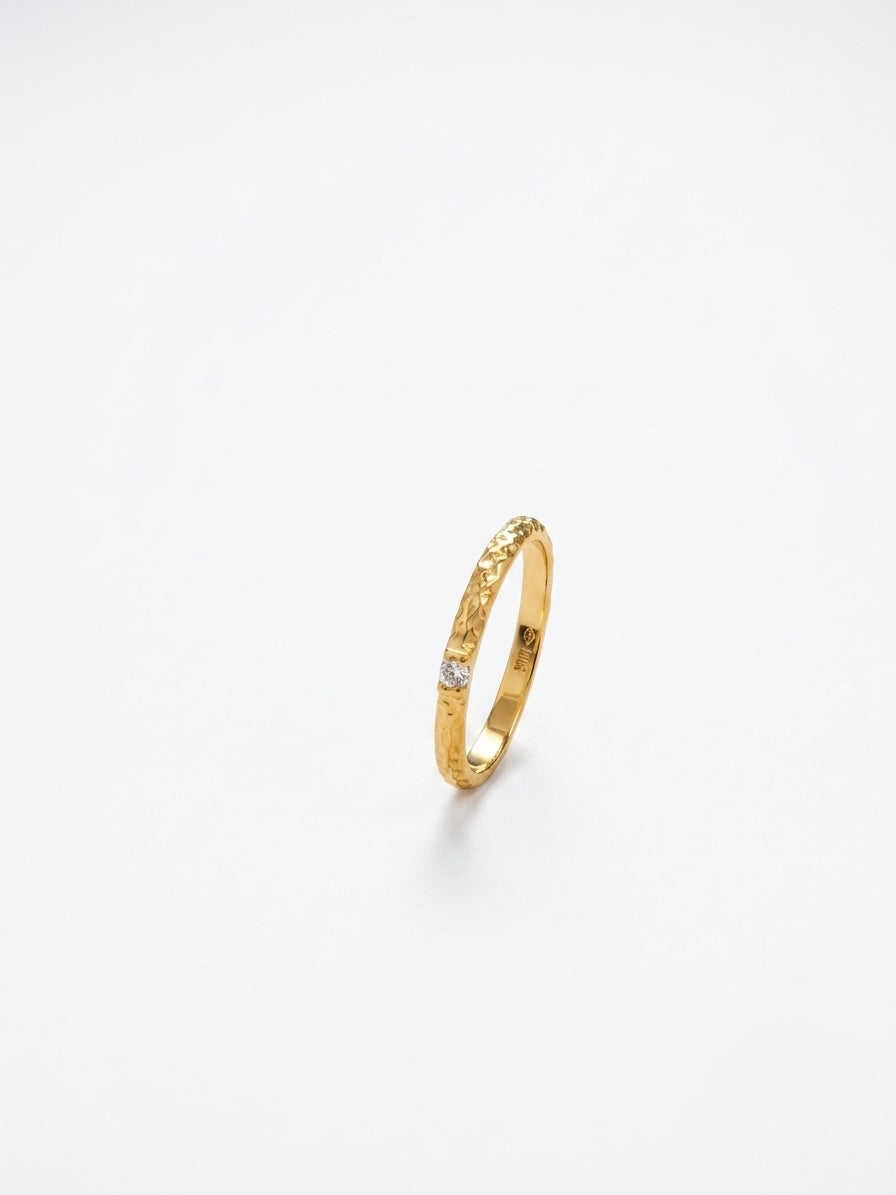 A delicate gold band ring with a textured, hammered finish and a small clear gemstone set near the front, photographed on a seamless white background.